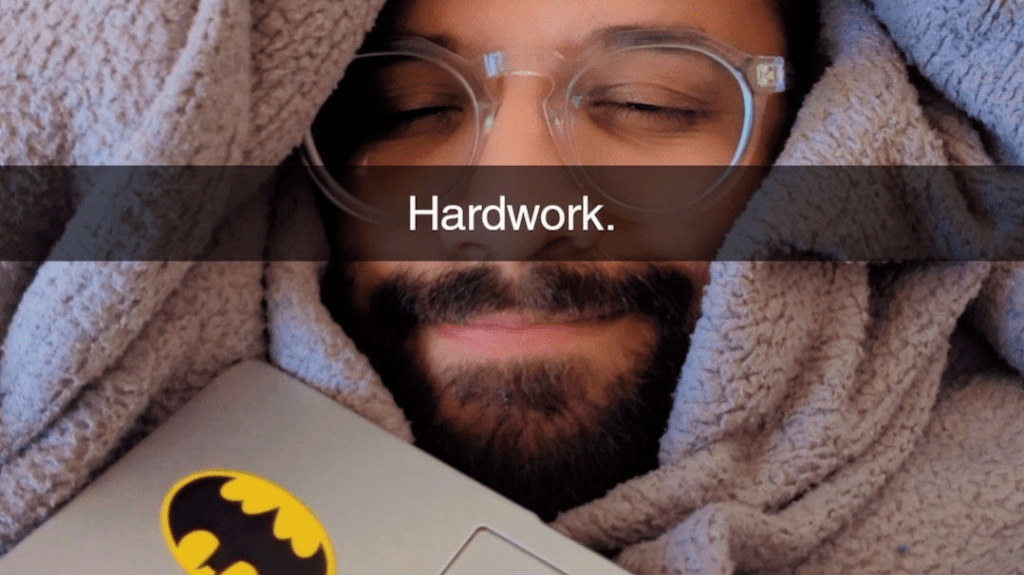 A person wrapped in a cozy blanket with glasses and a laptop featuring a Batman sticker, smiling peacefully, with the text 'Hardwork.' overlaying the image.